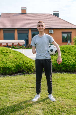 Full length young caucasian man with amputated arm, holds of soccer ball while standing on the lawn in the backyard of his home, disability rehabilitation and active lifestyle, vertical orientationの写真素材