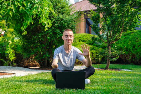 Young smiling guy with amputated arm sits on a lawn in backyard of his house and talking with friends via social network using laptopの写真素材