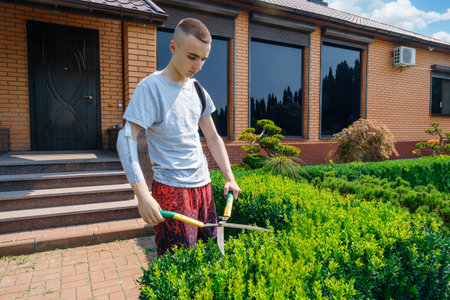 Young caucasian man with an amputated arm and a prosthesis is trimming bushes in a garden in his yard with large hedge shearsの写真素材