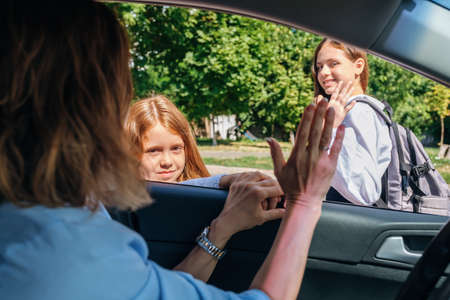Mother sitting inside the car and gesturing with hand goodbye and leaving her daughters in school uniform at school.の写真素材