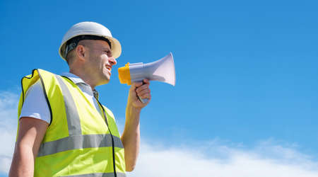 Builder, foreman or architect in a white helmet and a reflective vest shouts into a megaphone against a blue sky background, copy spaceの写真素材
