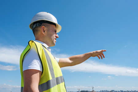 A builder, foreman or architect in a white helmet and a reflective vest shows with his hand on the construction against the blue sky background, copy spaceの写真素材