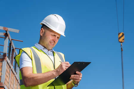 Mature man in white helmet stands on the roof, holds clipboard, inspects construction site and writing notes on blue sky background, copy space for textの写真素材