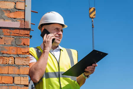 Portrait of mature man in white helmet at construction site, who checks the schedule while holding a clipboard and talking on a cell phone against a blue sky backgroundの写真素材