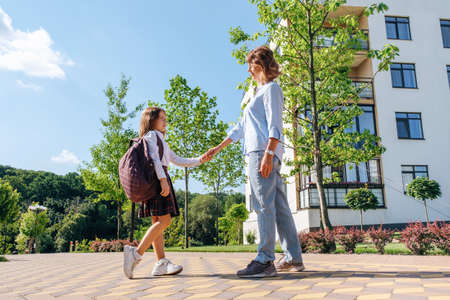 Back to school. Caucasian schoolgirl with a backpack and uniform holds her smiling mother by the hand in the street before going to school.の写真素材