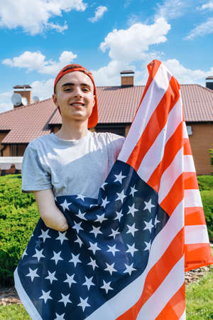 Happy young man with amputated arm looking at camera with smile and showing flag of United States while celebrating Independence Day in suburban yardの写真素材
