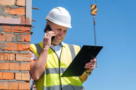 Happy male contractor smiling and reading data on clipboard while leaning on brick wall and answering phone call against blue sky on construction siteの写真素材