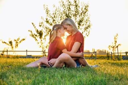 Side view of woman and girl in similar red t shirts touching foreheads and embracing each other, while sitting on green lawn against sundown skyの写真素材