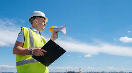 Builder, foreman or architect in a white helmet and a reflective vest shouts into a megaphone and holds a clipboard against a blue sky background, copy space, bannerの写真素材