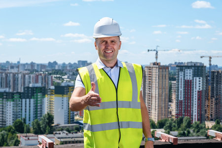 Adult man in hardhat and helmet smiling and holds out one hand for a handshake on the roof of a...