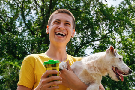 Young smiling caucasian man holds a Jack Russell terrier dog in his arms and drinks coffee from a disposable cup on a walk in the park, summertime.の写真素材