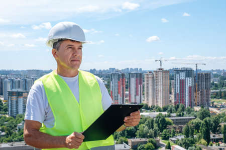 Portrait of mature man in white helmet at construction site who checks schedule while holding clipboard on urban background behind his backの写真素材