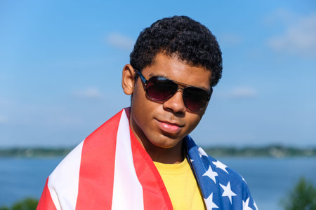 Portrait of young African American man in the sunglasses looking at camera and covered with American...