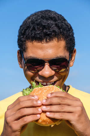 Hungry smiling African American man in sunglasses enjoying the taste of hamburger outdoor in summer. Concept of love to junk food diet, vertical orientationの写真素材