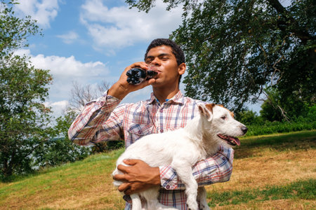 Young African American man holding Jack Russell terrier dog and drinking cola from bottle outdoors. Walking in park in summer, low angle viewの写真素材
