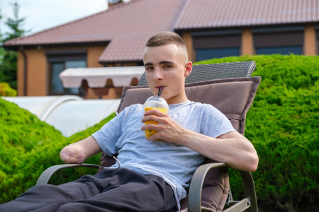 A young smiling male with an amputated arm lies on a sun lounger and drinks a cocktail, in the courtyard of his house on the lawnの写真素材
