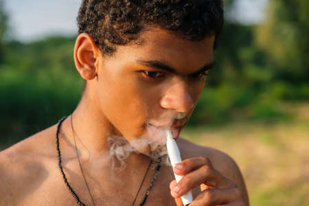 Close up face of young black african american man smoking electronic cigarette with e-liquid outdoors in summer, headshotの写真素材