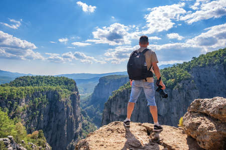 Full body back view of unrecognizable male hiker with backpack and photo camera in hand standing on edge of rocky cliff in highlandsの写真素材