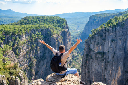 Back view of anonymous male hiker with backpack sitting on edge of rocky cliff with raised arms while admiring view of mountains in highlandの写真素材