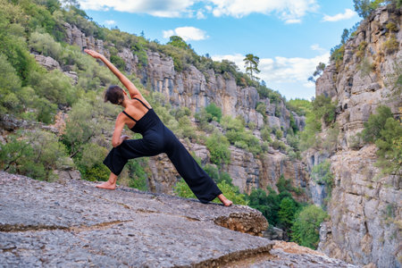 Back view full body of anonymous barefoot slender female practicing Virabhadrasana posture while standing on rocky cliff during yoga training in mountainous terrainの写真素材