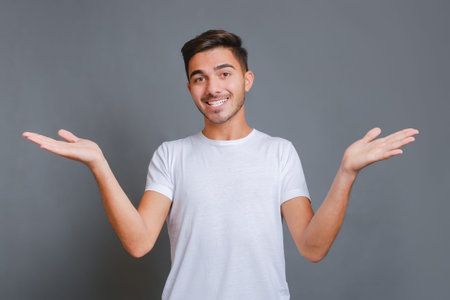 Young attractive smiling guy in white t-shirt making welcome gesture isolated on gray background