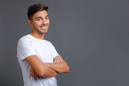 Handsome young unshaved man in white t-shirt with crossed arms looking at camera against gray wall...
