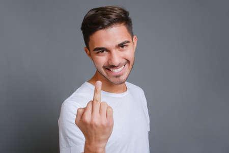 Close-up young smiling caucasian man wearing white t-shirt standing over isolated gray background and showing middle fingerの写真素材