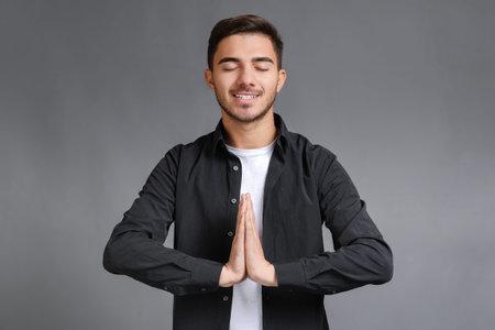 Bearded smiling young man with closed eyes wearing black shirt keeping hands namaste gesture and...