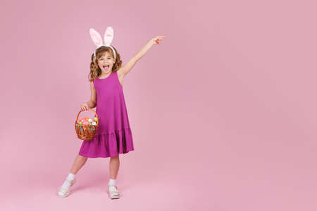 Full body little beautiful smiling girl in Easter bunny ears holds a basket with eggs on pink background and pointing away with hand. Copy space for textの写真素材