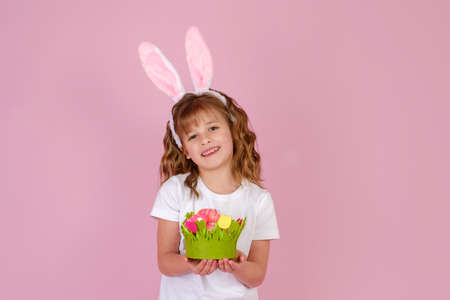 Little beautiful smiling girl in Easter bunny ears headband holds in hands Easter eggs on pink backgroundの写真素材