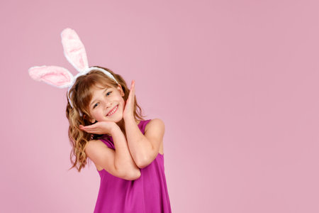Adorable little coquette with long curly hair in stylish fuchsia colored dress and bunny ears touching cheeks and smiling at camera, against ink background in studioの写真素材