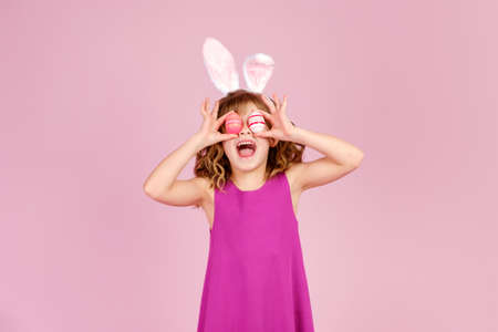 Adorable cheerful little girl with curly hair in fuchsia colored dress and bunny ear headband, smiling happily while covering eyes with eggs during Easter celebrationの写真素材