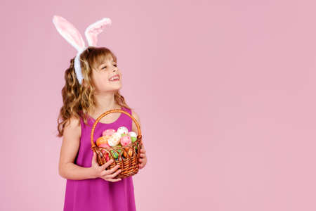 Cute positive little girl with curly hair in bright dress and bunny ears, smiling happily and looking away from the camera while holding wicker basket with colorful decorated eggs on a pink backgroundの写真素材