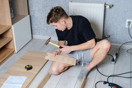 Serious young guy using hammer for assembling wooden furniture in room while sits on floor at home and working with detailsの写真素材