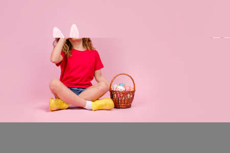 Little beautiful smiling girl in Easter bunny ears headband holds in hand a basket with Easter eggs,...