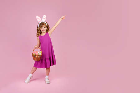 Full body little beautiful smiling girl in Easter bunny ears holds a basket with eggs on pink background and pointing away with hand. Copy space for textの写真素材