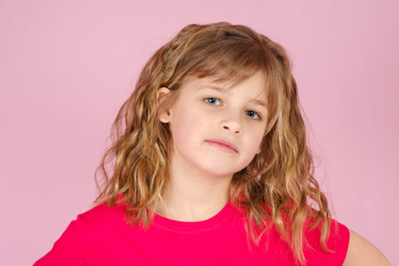 Close-up adorable little girl with long curly hair in red colored t-shirt looking at camera, against...
