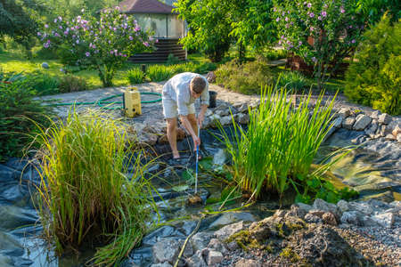 Young caucasian guy cleans artificial garden pond bottom with landing net from mud, sludge and water plants. Spring and summer pond care workの写真素材