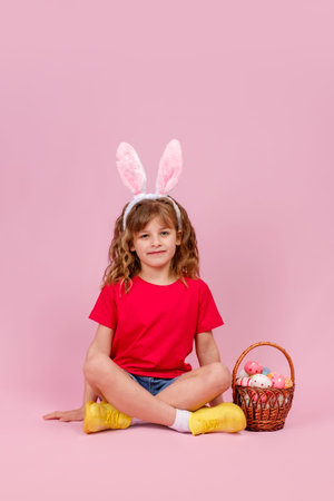 Little beautiful smiling girl in Easter bunny ears headband sitting on the floor near basket with...
