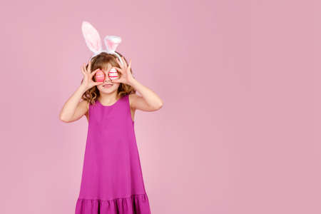 Adorable smiling little girl with curly hair in fuchsia colored dress and bunny ear headband, smiling happily while covering eyes with eggs during Easter celebration, copy spaceの写真素材