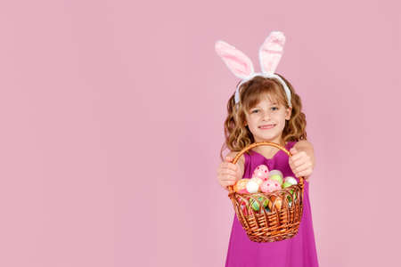 Adorable little girl with blond curly hair in stylish dress and bunny ears, smiling and looking at camera while demonstrating wicker basket full of colorful Easter eggs, copy spaceの写真素材