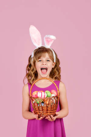 Adorable little girl with curly hair in stylish dress and bunny ears, smiling and looking at camera while demonstrating wicker basket full of colorful Easter eggs, vertical orientationの写真素材