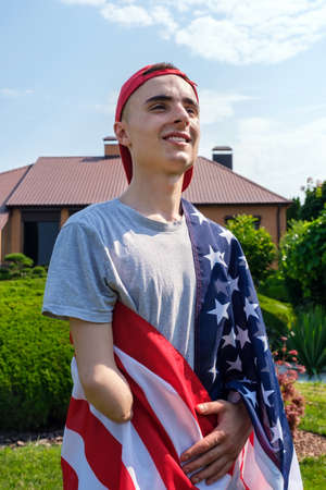 Happy young man with amputated arm with smile, looking away and covered with flag of United States while celebrating Independence Day in suburban yardの写真素材