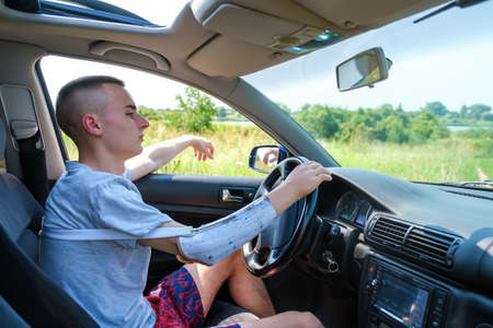 Young caucasian guy with amputated arm and prosthesis drives a car on a sunny day in summer. Side view inside of cabinの写真素材