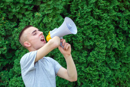 Young caucasian male with amputated arm shouting into a megaphone while standing against green vegetation backgroundの写真素材
