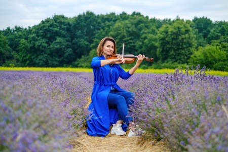 Full body adult woman violinist playing violin and sitting on summer lavender field, romantic musician in blue dress enjoying walking on natureの写真素材