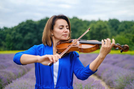 Close-up adult caucasian woman violinist playing violin on summer lavender field, romantic musician in blue dress enjoying walking on natureの写真素材