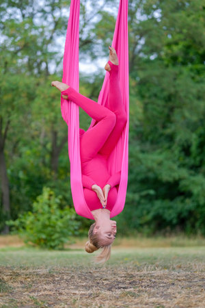 Beautiful young woman practices aerial yoga in a pink hammock hanging on a tree in the park. A woman practices anti-gravity yoga outdoors. Vertical orientationの写真素材