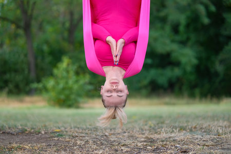 Beautiful young woman practices aerial yoga in a pink hammock hanging on a tree in the park. A woman practices yoga outdoors.の写真素材