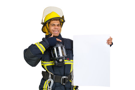 Young smiling African American fireman wearing uniform and helmet looking to the camera and holding blank paper sheet in hand and finger pointing. Copy space for text, isolated on white backgroundの写真素材